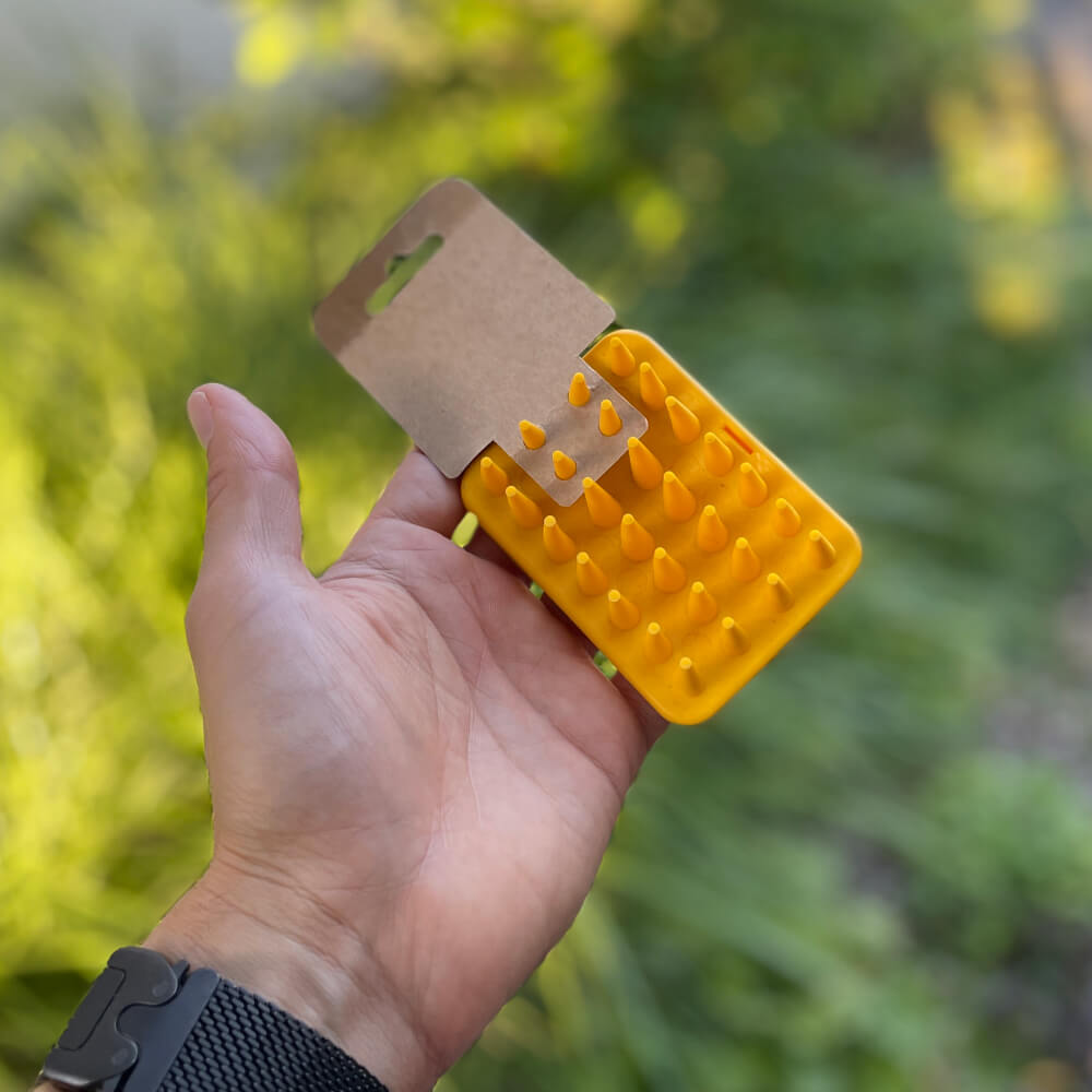 Żółta szczotka dla kota Fiboo prezentowana od strony tylnej z widoczną kartonową etykietą produktu. Ergonomiczna szczotka wykonana jest z elastycznego tworzywa, a w tle znajduje się rozmyty ogród.